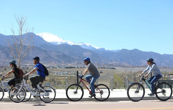 A group of four bikes bike down the road with Pikes Peak visible in the background.